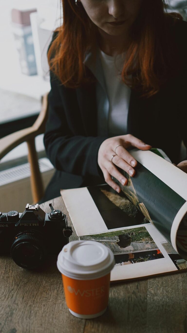 Redheaded woman sitting in a café, flipping through a photo album with a camera and coffee cup nearby.
